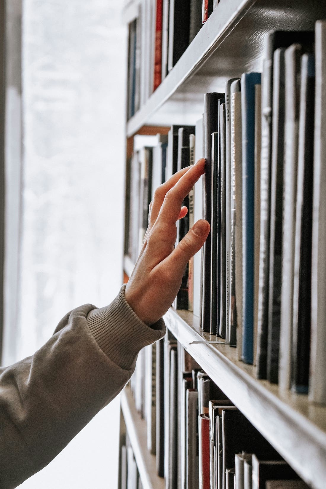 A hand reaching out to pluck a book off a library book shelf, the spines unreadable.